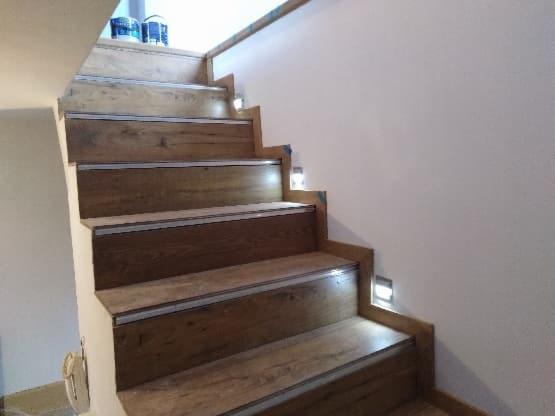 Drewniane schody z oświetleniem LED wbudowanym w ścianę, widoczne puszki z farbą na górnym stopniu