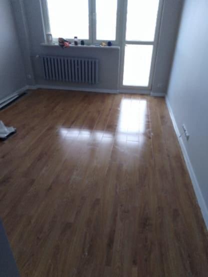 Wykończona podłoga z paneli w jasnym odcieniu drewna, widoczny grzejnik pod oknem i szare ściany w pustym pokoju.