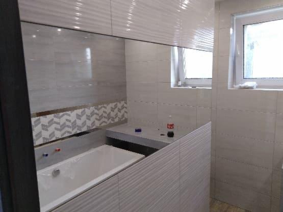 Nowoczesna łazienka z wanną obudowaną płytkami imitującymi drewno, szare płytki ścienne z subtelnym wzorem jodełki, okno z białymi ramami, butelka Coca-Coli i niebieski przedmiot na blacie.