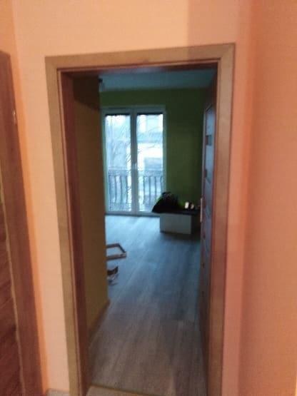 Widok przez odnowione drzwi w Chorzowie na pokój z nową podłogą i świeżo pomalowanymi ścianami w odcieniach zieleni.