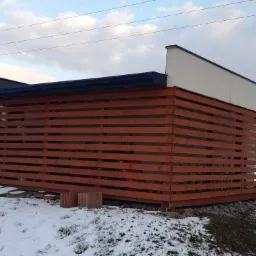 Nowoczesna altana z poziomych desek w kolorze brązu, z płaskim dachem i białymi ścianami, otoczona śniegiem w zimowym krajobrazie podmiejskim.