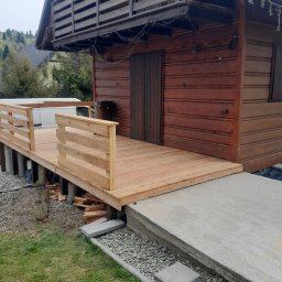 F.H.U CIESIELSKI PIOTR - Nowy, jasny taras drewniany z balustradą przy domku z bali, widok z zewnątrz. Elementy betonowe i kamienne na gruncie. W tle zieleń i zabudowa.