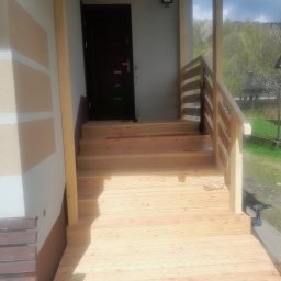F.H.U CIESIELSKI PIOTR - Drewniany taras z balustradą i schodami przed wejściem do domu. Widoczne świeże deski i narzędzie pracy. W tle zieleń i zabudowania.