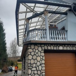 F.H.U CIESIELSKI PIOTR - Zadaszony balkon z balustradą i kolumnami, wykończony kamieniem. Widoczna konstrukcja z przezroczystego tworzywa. Na dole garaż z brązową bramą segmentową.