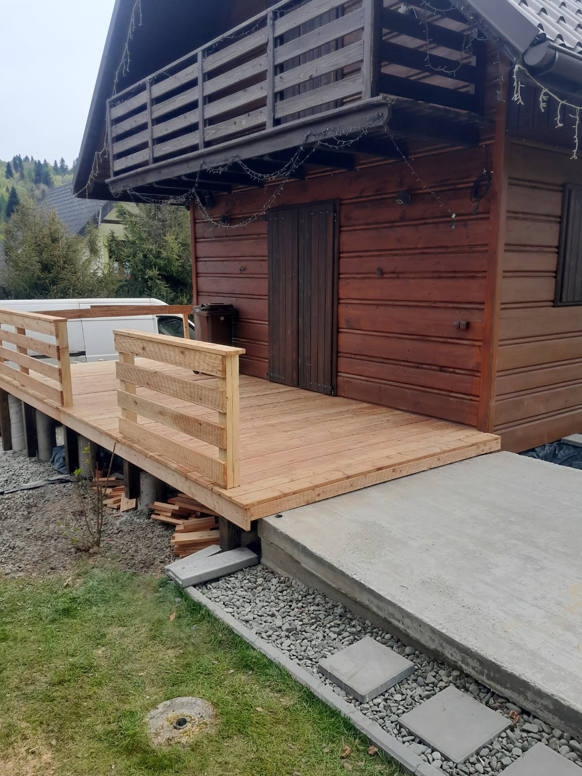 Nowy, jasny taras drewniany z balustradą przy domku z bali, widok z zewnątrz. Elementy betonowe i kamienne na gruncie. W tle zieleń i zabudowa.