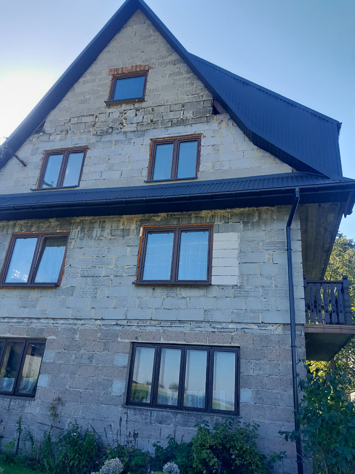 Dwupiętrowy dom w trakcie budowy lub remontu z zamontowanymi oknami i częściowo wykończonym dachem z blachy w kolorze antracytowym, widok z dołu.