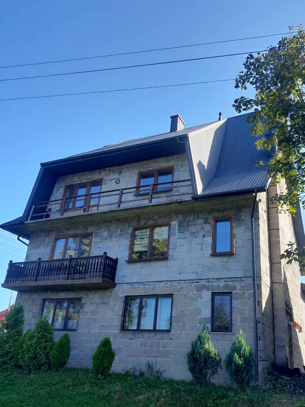 Dwupiętrowy dom z szarej cegły z ciemnym, metalowym dachem i drewnianymi balkonami w słoneczny dzień. Widoczne rynny i komin.