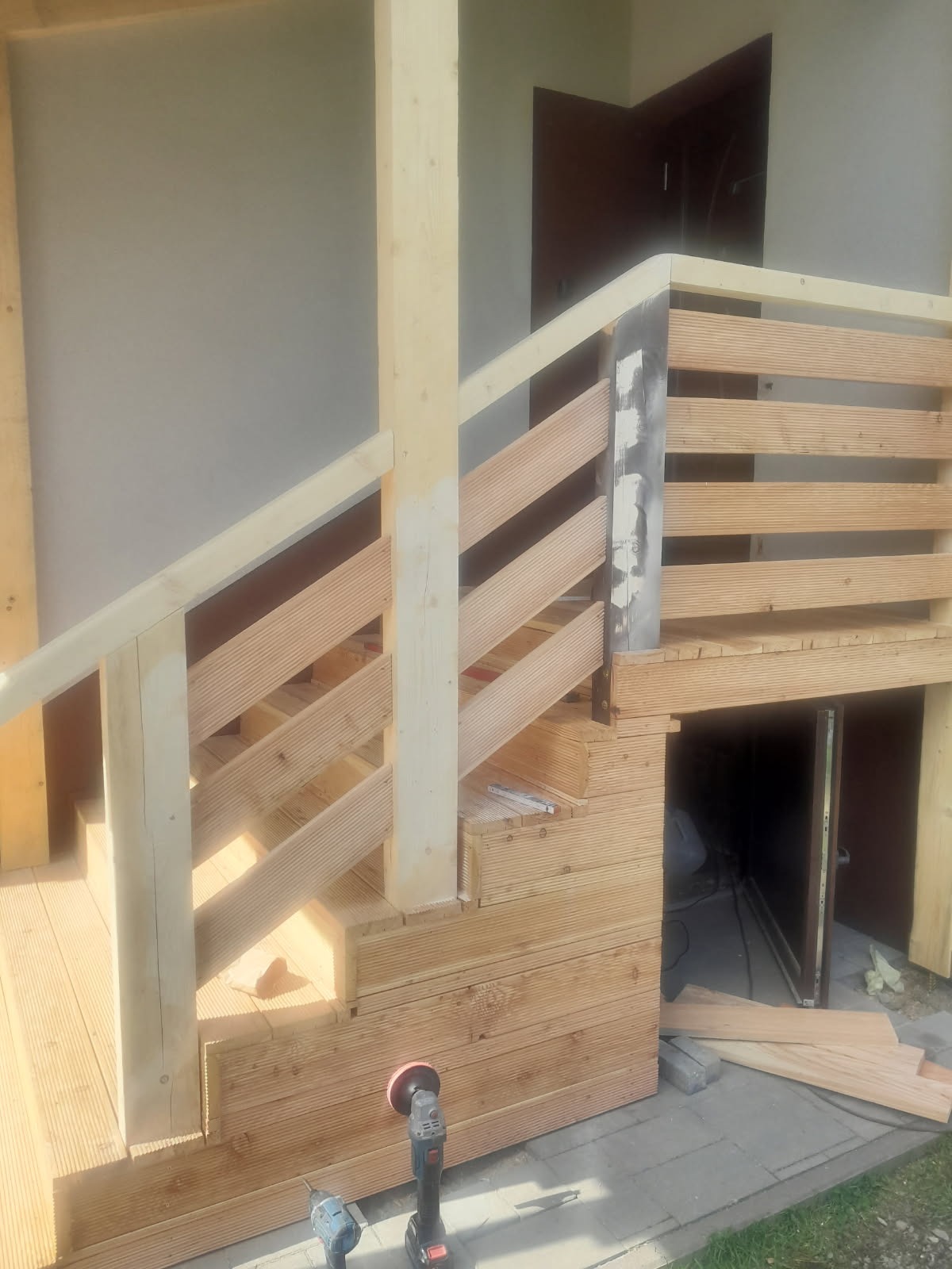 Drewniany taras w trakcie budowy z widocznymi schodami, balustradą i narzędziami. Jasne drewno, częściowo wykończone, z detalami montażowymi.