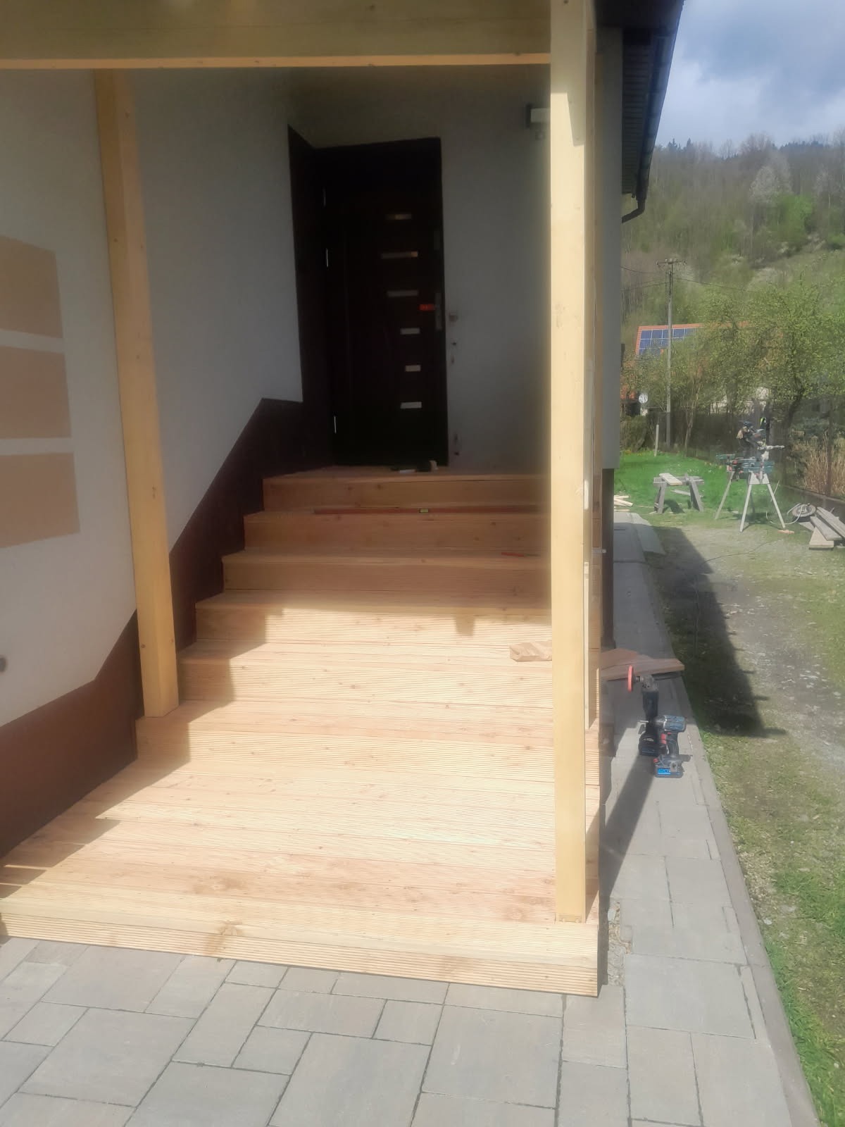 Drewniany taras z schodami w trakcie budowy. Widoczne narzędzia, elementy konstrukcji i krajobraz w tle. Świeże drewno, jasne oświetlenie.