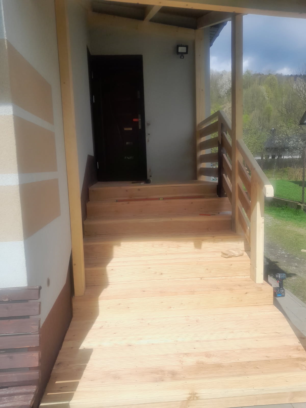 Drewniany taras z balustradą i schodami przed wejściem do domu. Widoczne świeże deski i narzędzie pracy. W tle zieleń i zabudowania.