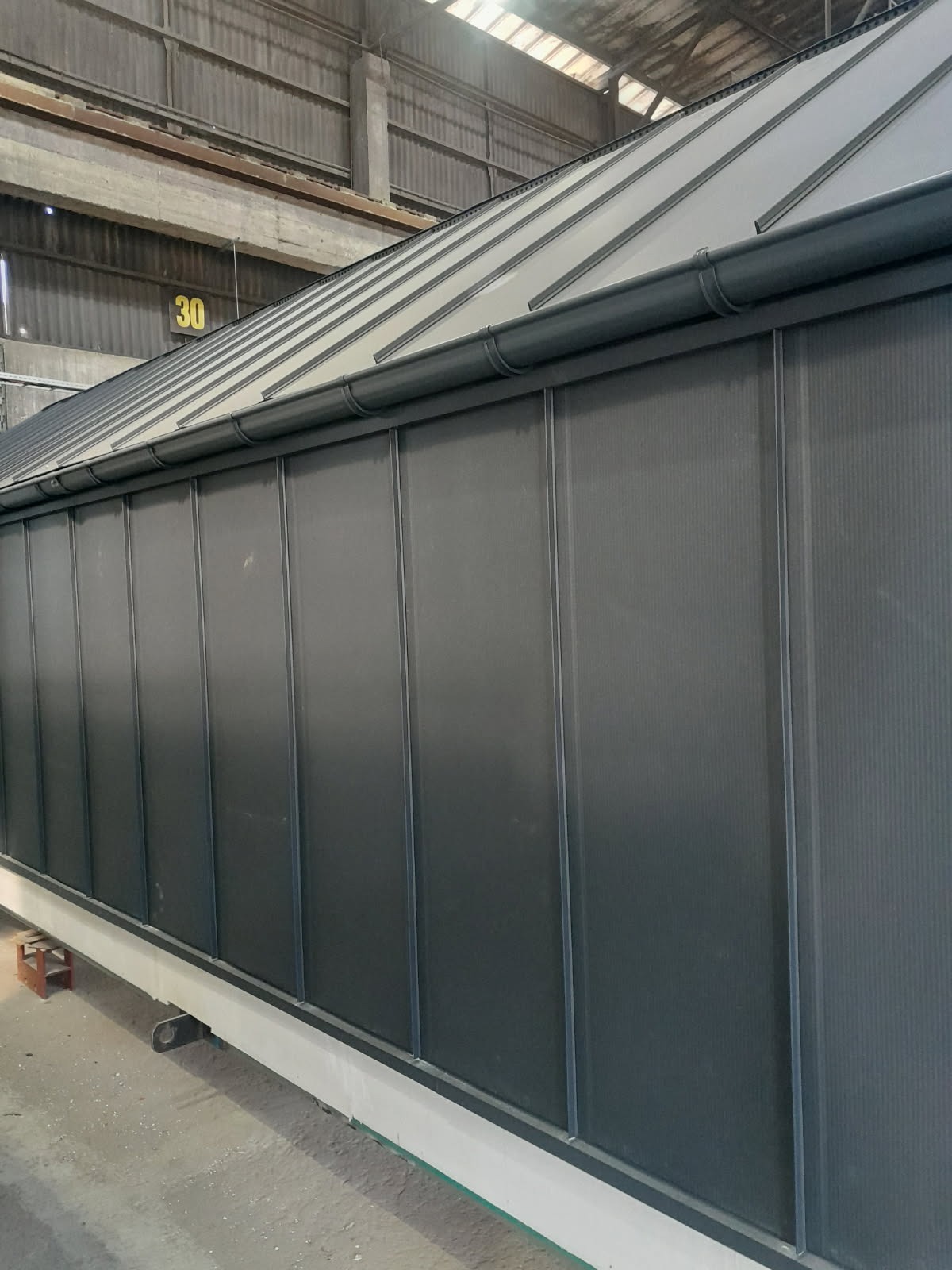 Szara elewacja z blachy trapezowej z rynnami w hali produkcyjnej, widok z dołu. Element konstrukcji z numerem 30 w tle.