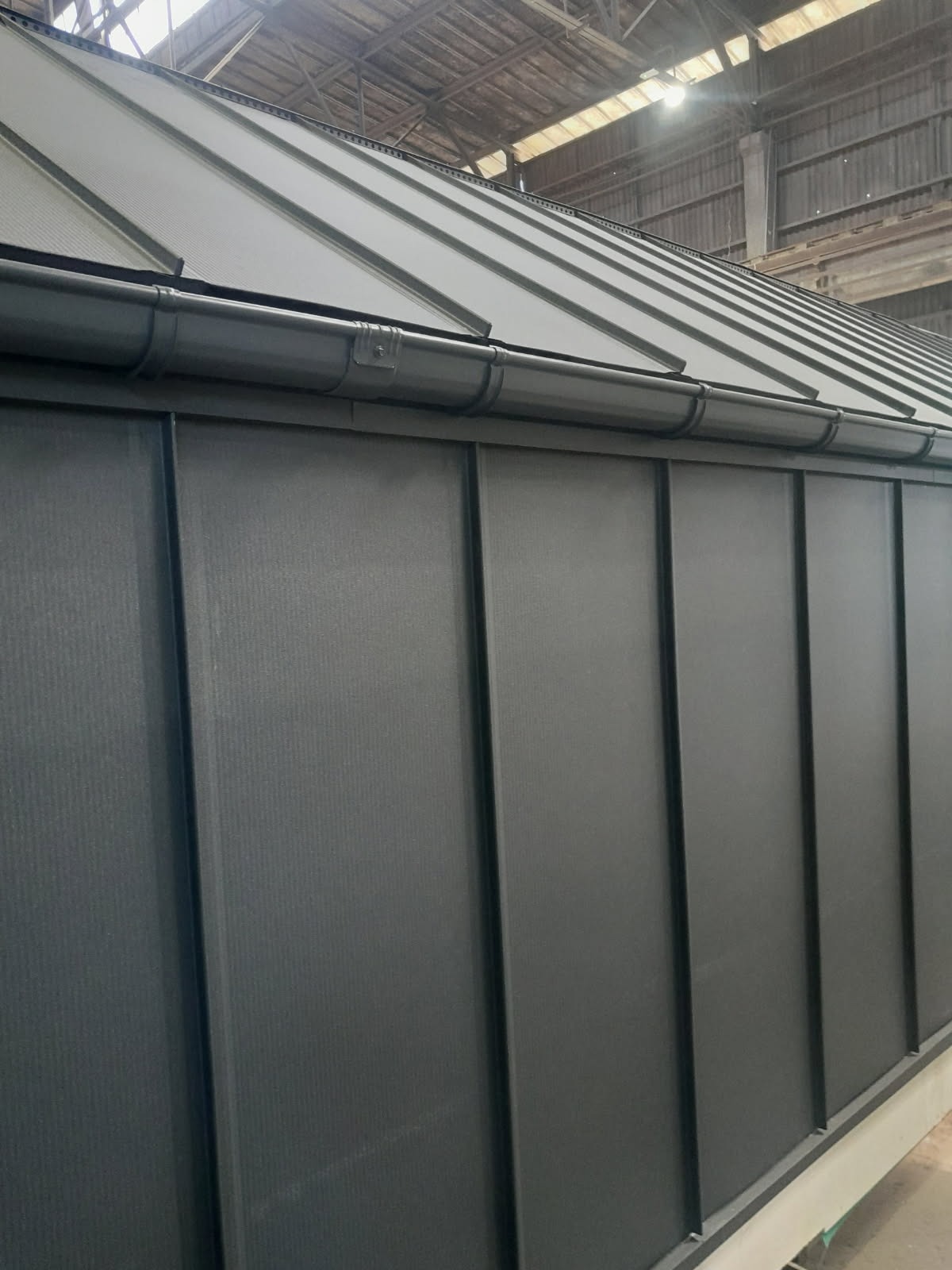 Szara blacha na dachu i elewacji, widoczne rynny. Ujęcie z dołu, podkreślające regularne linie i nowoczesny wygląd. Hala w tle.