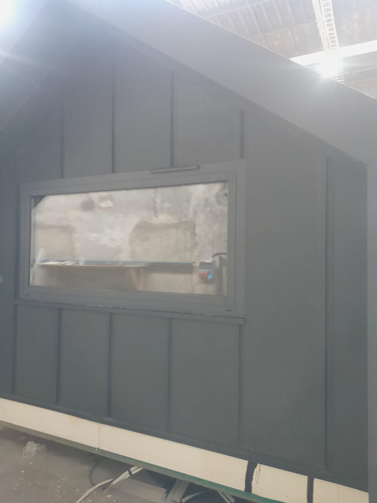 Szara konstrukcja z oknem, przypominająca domek narzędziowy lub fragment budynku z blachy trapezowej. Widoczne łączenia blachy i rama okienna.