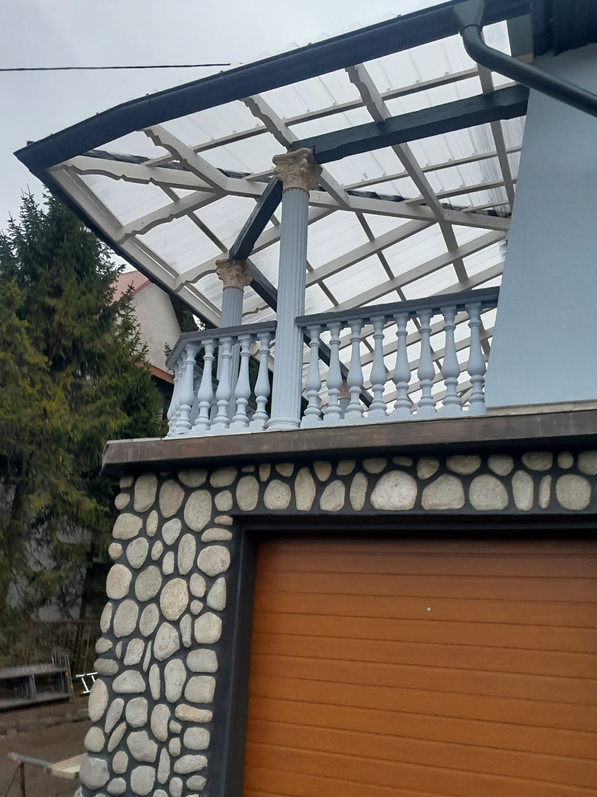 Zadaszenie tarasu z kolumnami i balustradą, wykończone kamieniem rzecznym nad bramą garażową. Widok z dołu, pochmurne niebo w tle.