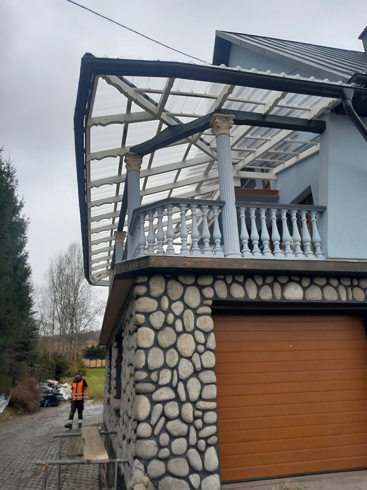 Zadaszony balkon z balustradą i kolumnami, wykończony kamieniem. Widoczna konstrukcja z przezroczystego tworzywa. Na dole garaż z brązową bramą segmentową.