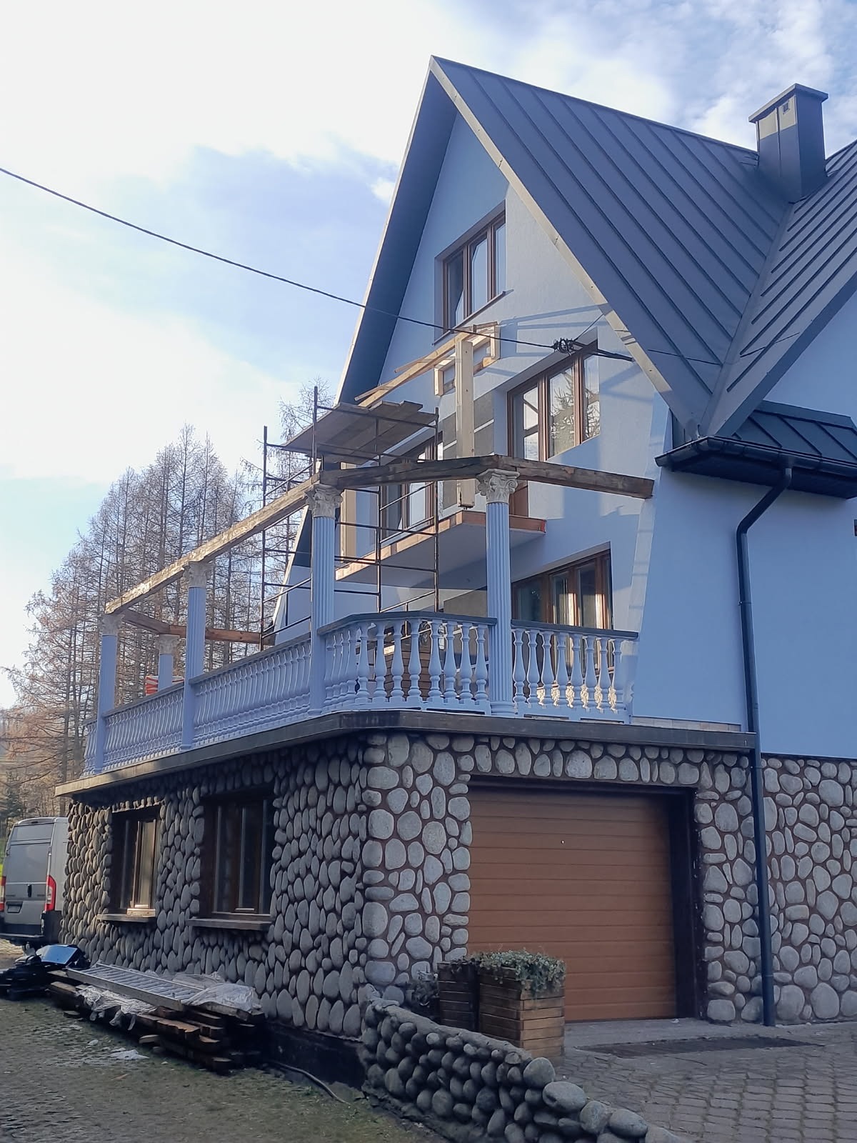 Dom z balkonem w trakcie budowy/remontu; widoczne rusztowania i elementy konstrukcyjne. Dach pokryty blachą, elewacja w odcieniach szarości i błękitu, kamienna podstawa.