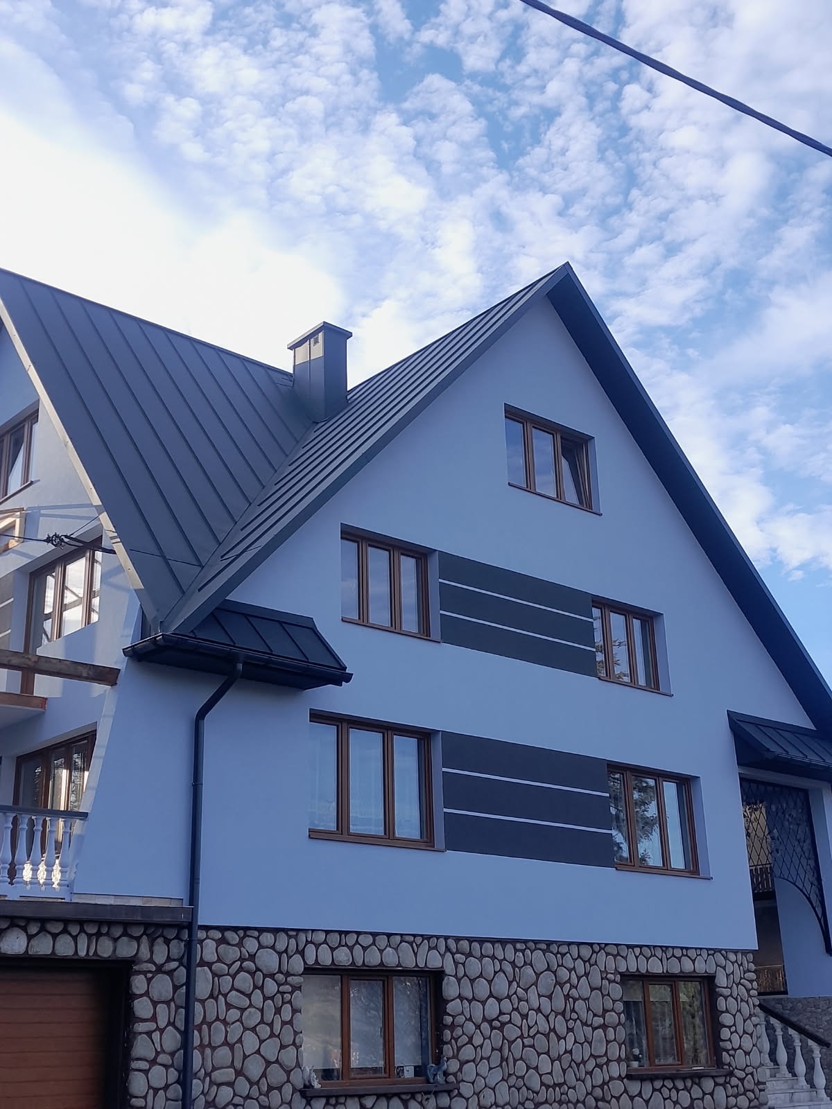 Dwupiętrowy dom z ciemnoszarym dachem dwuspadowym i jasnoniebieską elewacją z dekoracyjnymi szarymi pasami i kamienną podmurówką, uchwycony pod kątem z dołu.