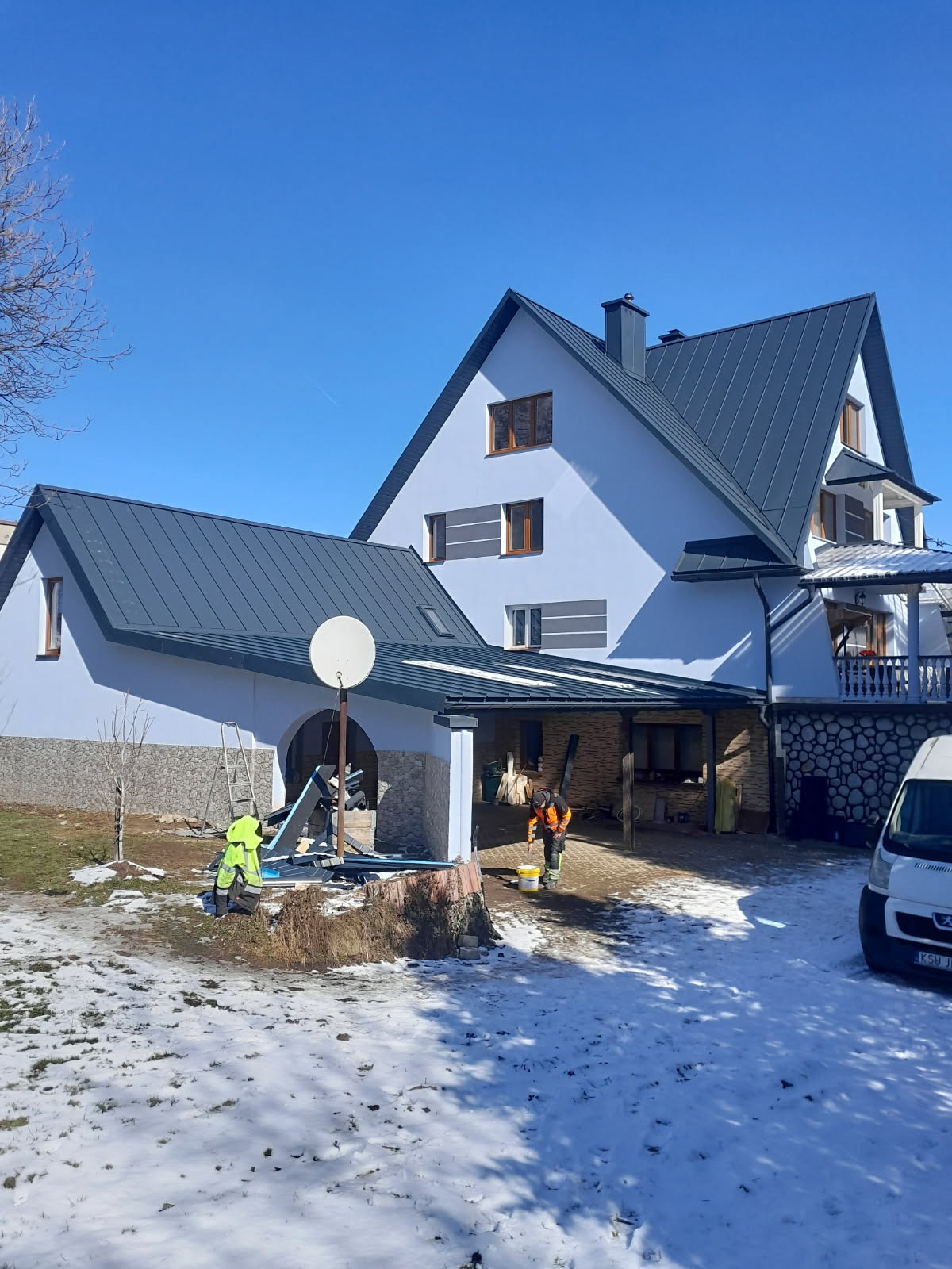 Dwóch pracowników w odblaskowych ubraniach pracuje przy domu z ciemnoszarym dachem w zimowej scenerii. Jeden maluje element konstrukcyjny, drugi przygotowuje materiały.