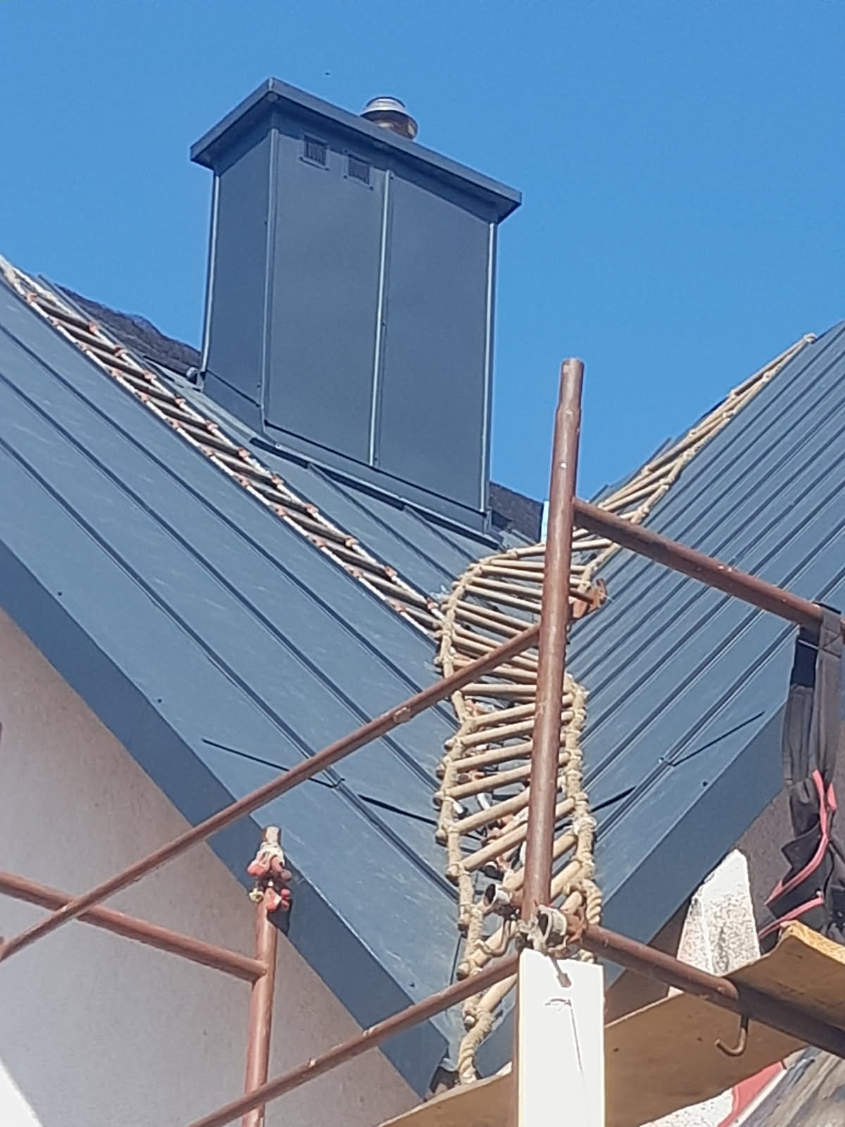 Ciemnoszara blacha na dachu z kominem i drabinką dekarską, widziana z rusztowania. Dachówka ułożona w regularne pasy, na tle błękitnego nieba.