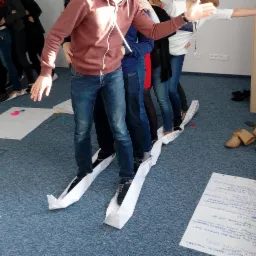 Grupa osób w jeansach i butach stoi w rzędzie, balansując na długim, poskładanym pasku papieru na podłodze, podczas ćwiczenia zespołowego.