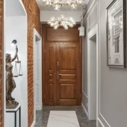Wnętrze przedpokoju z elementami dekoracyjnymi: statuetka Temidy na postumencie, ceglana ściana z łukiem, marmurowa posadzka z białym pasem, drewniane drzwi wejściowe, dwie okrągłe lampy sufitowe...