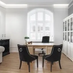 Jasne, przestronne biuro w Gdańsku z biurkiem, dwoma czarnymi krzesłami z ozdobnymi kółkami na oparciach, białymi regałami z segregatorami i komodą z dekoracjami. Duże okno z żaluzjami w tle.