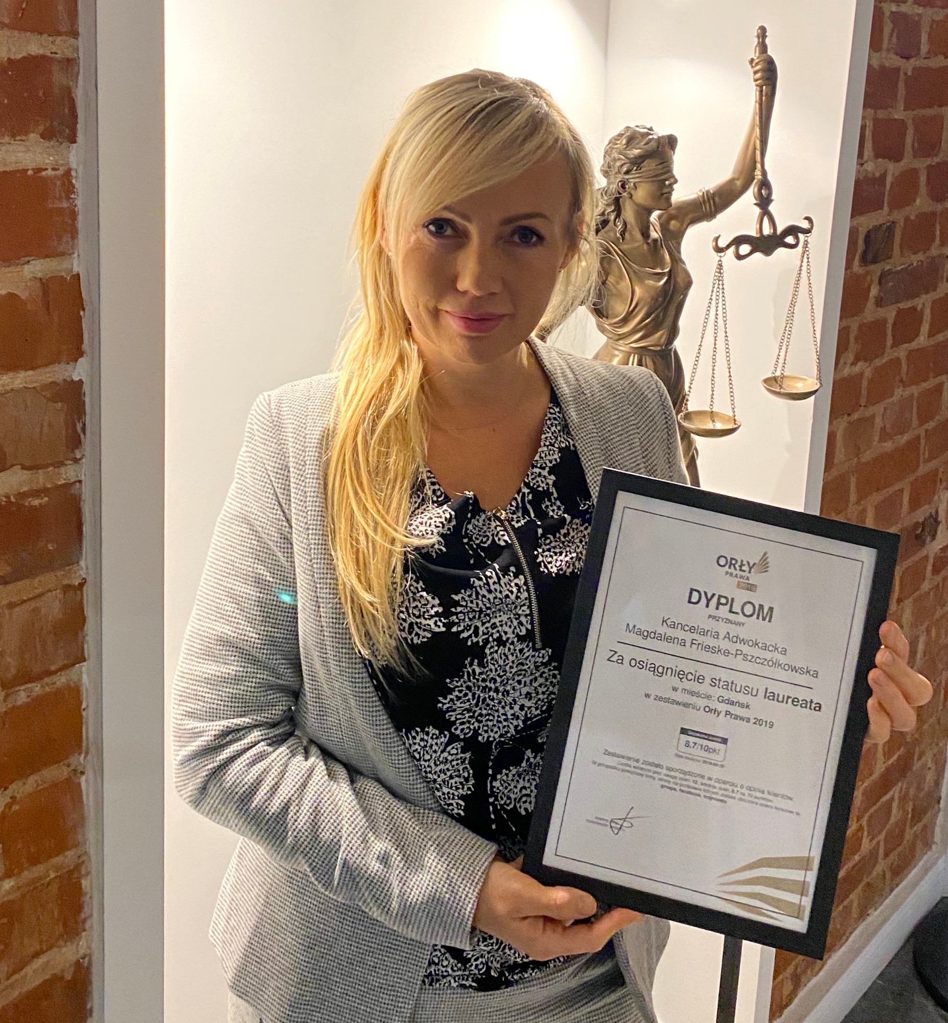 Kobieta w szarym żakiecie prezentuje dyplom 'Orły Prawa 2019' dla Kancelarii Adwokackiej Magdaleny Frieske-Pszczółkowskiej, w tle statuetka Temidy i fragment ściany z cegły.