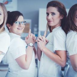 Vidium Medica - Zespół medyczny w białych uniformach, uśmiechnięte kobiety z uniesionymi palcami wskazującymi, jedna z nich w okularach i z zegarkiem na ręce.