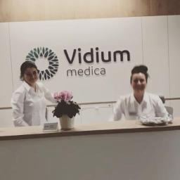 Vidium Medica - Recepcja kliniki Vidium Medica z dwiema pracownicami w białych uniformach, dekorowana doniczką z różowymi kwiatami.