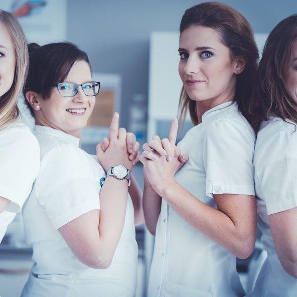 Zespół medyczny w białych uniformach, uśmiechnięte kobiety z uniesionymi palcami wskazującymi, jedna z nich w okularach i z zegarkiem na ręce.