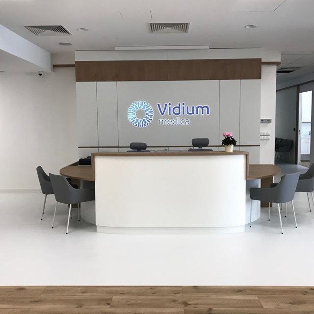 Jasne, minimalistyczne wnętrze recepcji kliniki medycznej z logo Vidium Medica na ścianie, okrągły blat i szare krzesła dla pacjentów.