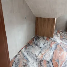 Wykończone wnętrze poddasza z szarymi płytkami na ścianach i wzorzystą, marmurkową podłogą w odcieniach szarości, bieli i brązu, widoczny fragment drewnianej zabudowy w rogu pomieszczenia...