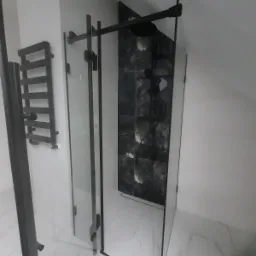 Nowoczesna kabina prysznicowa z czarnymi profilami, marmurowe płytki na ścianie pod prysznicem i biała podłoga z efektem marmuru, widoczny grzejnik drabinkowy.