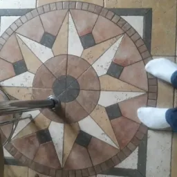 Podłoga wyłożona płytkami z wzorem rozety w odcieniach beżu, brązu i szarości, widoczne stopy w białych skarpetkach i fragment metalowej nogi krzesła.