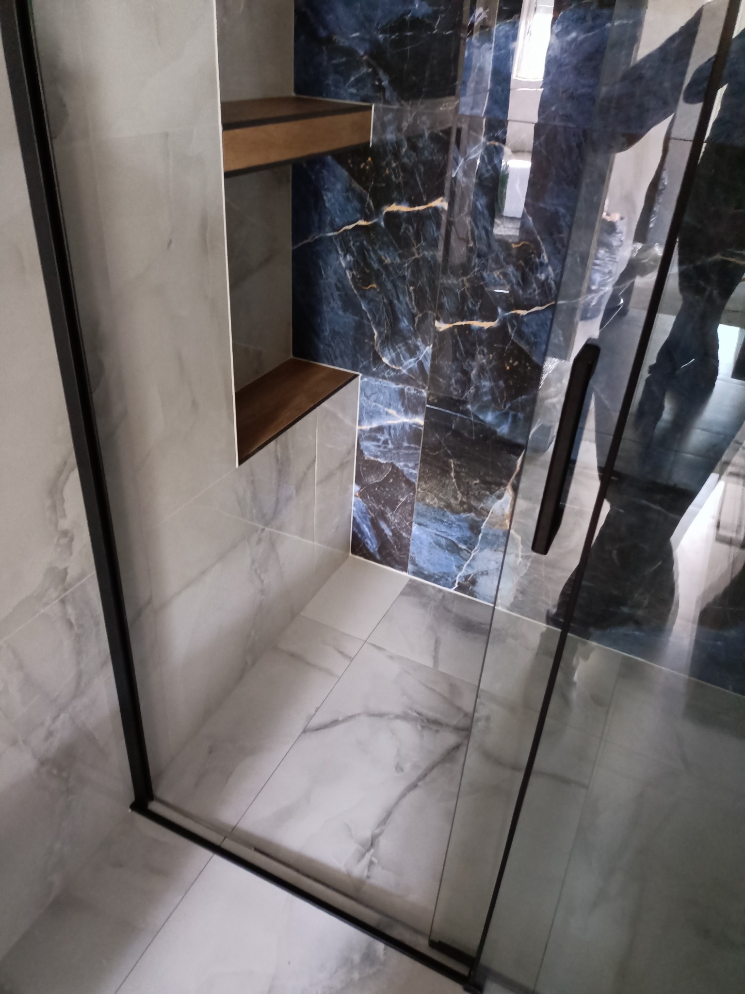 Wykończona łazienka z kabiną prysznicową typu walk-in z czarnymi profilami, marmurową glazurą w odcieniach szarości i błękitu oraz drewnianymi półkami wnękowymi.
