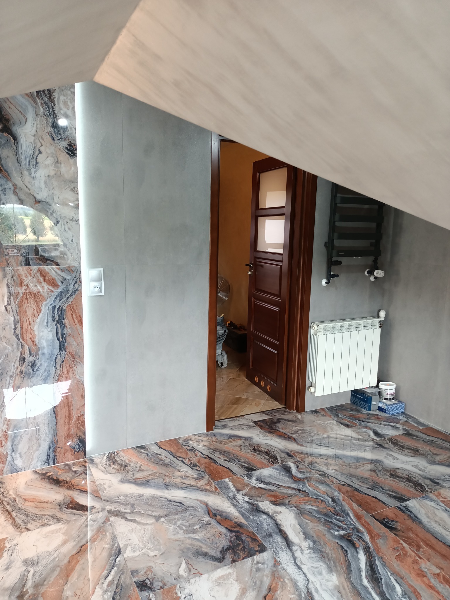 Ukończona łazienka na poddaszu z marmurową płytką podłogową i ścienną, widoczny otwarty drewniany drzwi, grzejnik i akcesoria malarskie.