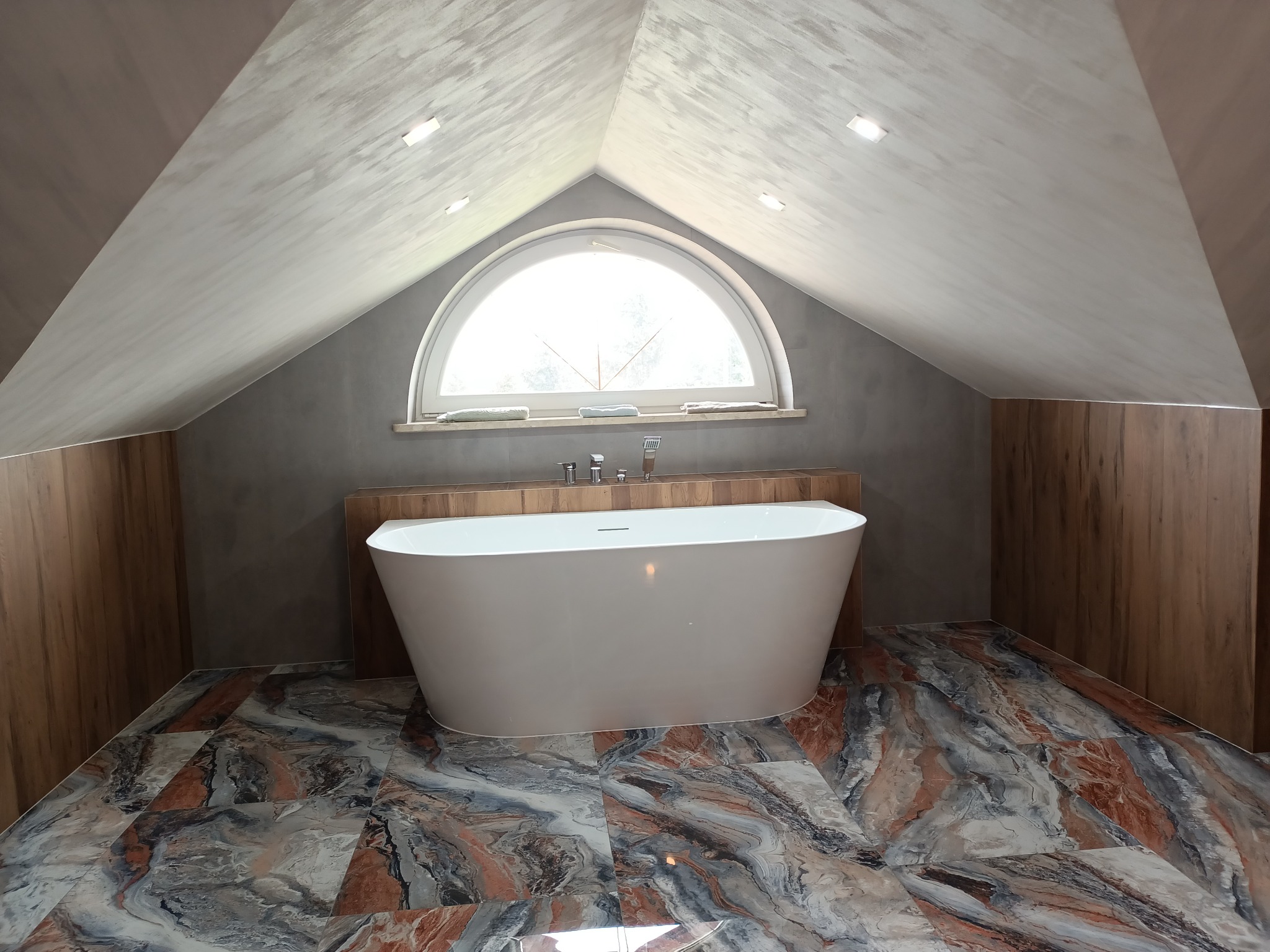 Nowoczesna wanna wolnostojąca w łazience na poddaszu z oknem w kształcie półkola i marmurową posadzką w odcieniach szarości, błękitu i brązu.