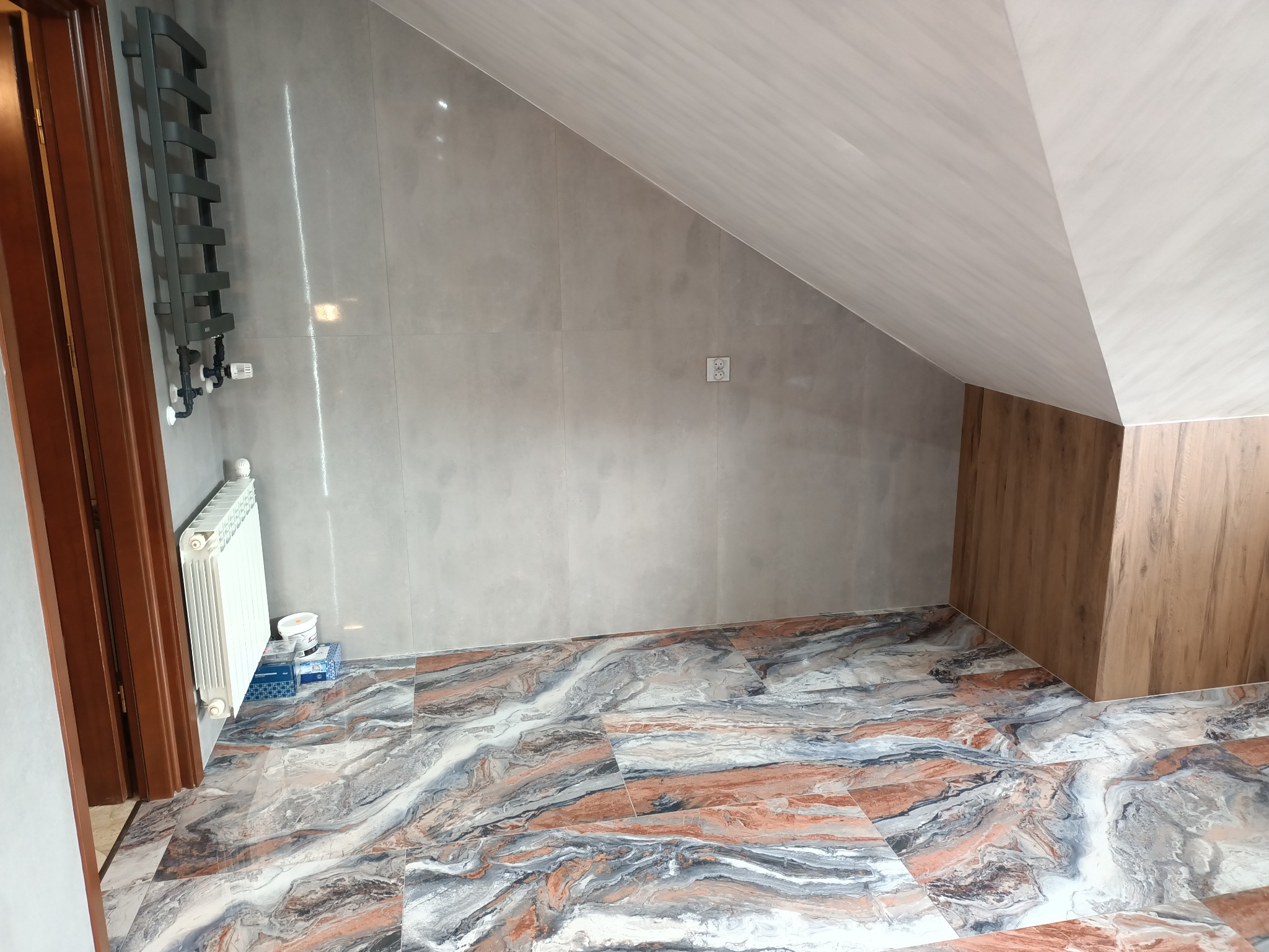 Ukończona łazienka na poddaszu z szarymi płytkami ściennymi, podłogą w marmurkowy wzór, grzejnikiem drabinkowym i grzejnikiem żeberkowym. Widoczne puszki z farbą i materiały budowlane.