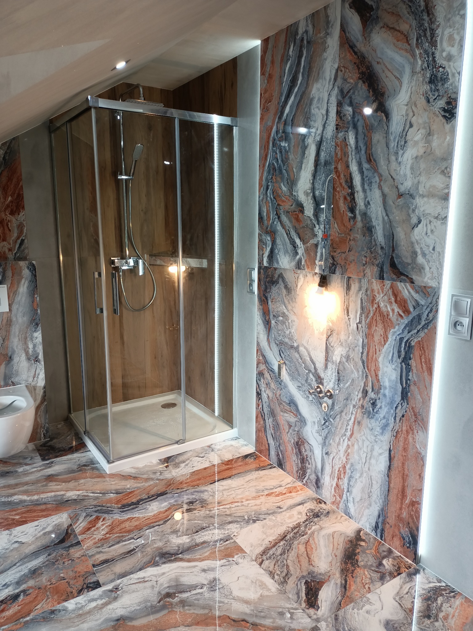 Kabina prysznicowa z chromowaną armaturą na tle ściany z płytek imitujących marmur w odcieniach bieli, szarości i rdzawej czerwieni, z podświetleniem LED po prawej stronie i płytkami podłogowymi...