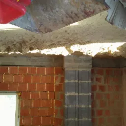 Wnętrze w trakcie prac murarskich: ściany z czerwonej cegły i szarego betonu, surowy sufit z widoczną konstrukcją i wystającymi elementami, okno z białymi ramami, nieporządek budowlany.