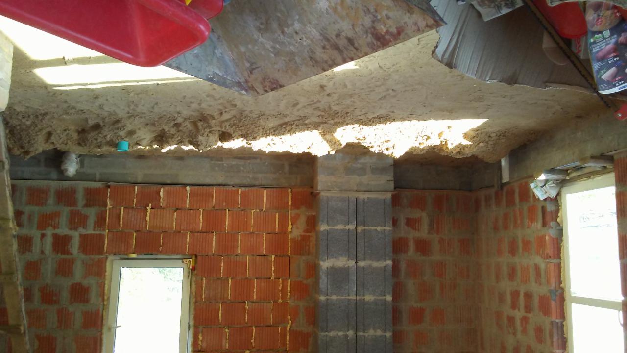 Wnętrze w trakcie prac murarskich: ściany z czerwonej cegły i szarego betonu, surowy sufit z widoczną konstrukcją i wystającymi elementami, okno z białymi ramami, nieporządek budowlany.