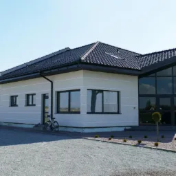 Nowoczesny budynek z dużymi przeszkleniami i czarną dachówką, rower oparty o ścianę, żwirowe podłoże.