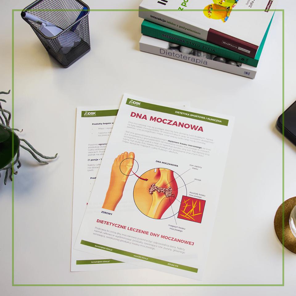 Ujęcie z góry na biurko z broszurami informacyjnymi o dnie moczanowej, książkami o dietoterapii i interakcjach leków, metalowym koszykiem na długopisy i rośliną w doniczce.