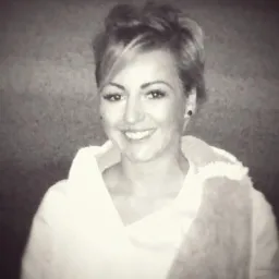 Portret uśmiechniętej kobiety z krótkimi włosami, ubrana w białą bluzkę z futrzanym kołnierzem, na rozmytym, ciemnym tle.