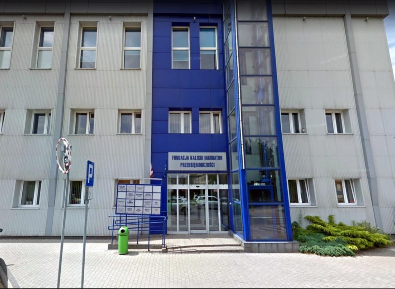 Budynek Fundacji Kaliski Inkubator Przedsiębiorczości z niebieskim akcentem, windą zewnętrzną i zielonym koszem na śmieci przed wejściem.