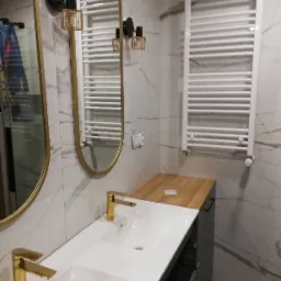 Wykończona łazienka z dwoma umywalkami nablatowymi z bateriami w kolorze złotym, podwieszaną szafką, dwoma owalnymi lustrami w złotych ramach, grzejnikiem drabinkowym i marmurowymi płytkami...