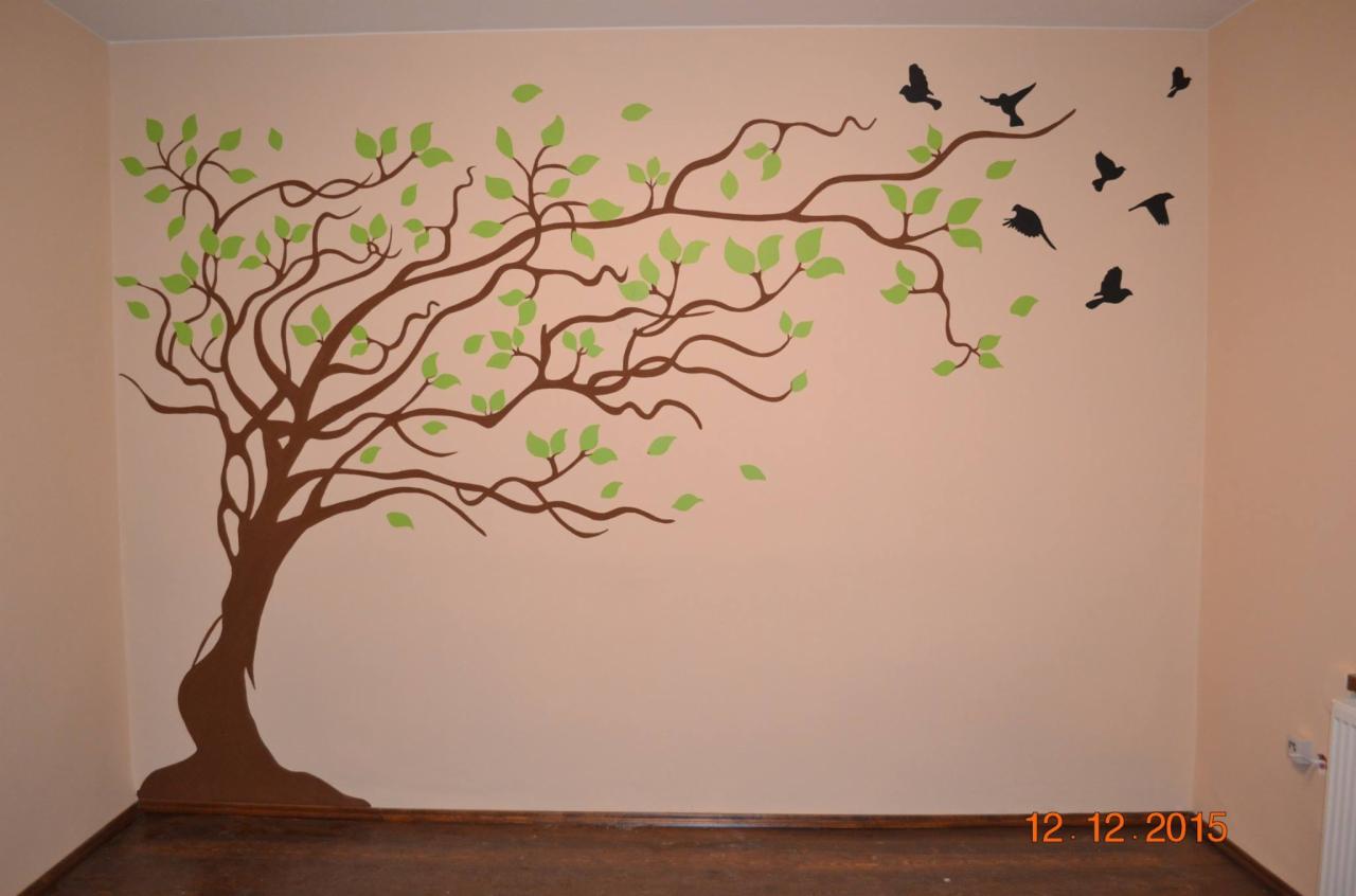Ściana pokoju z ręcznie malowanym motywem drzewa z zielonymi liśćmi i odlatującymi czarnymi ptakami.