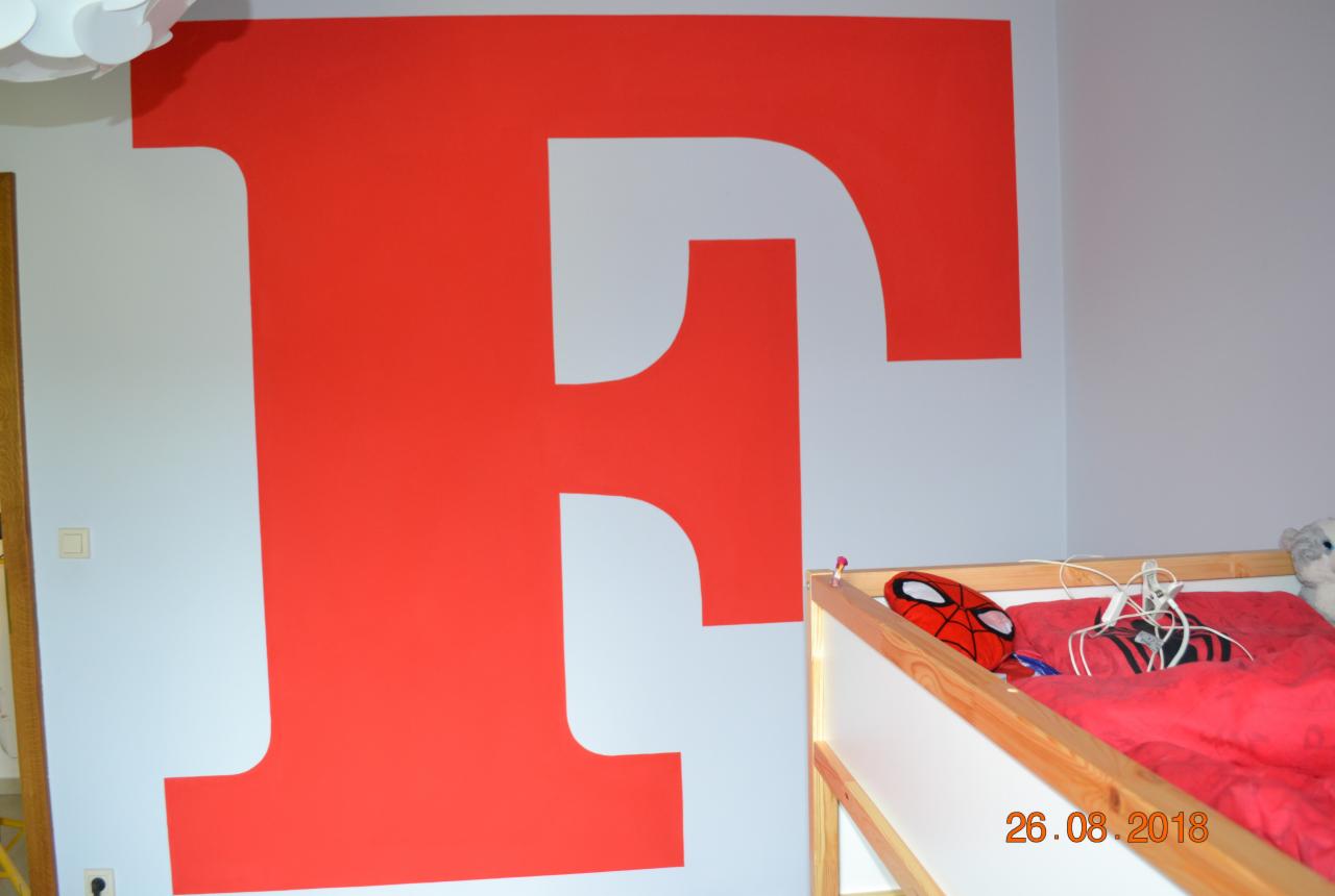 Duża, czerwona litera F namalowana na szarej ścianie w pokoju dziecięcym, widoczny fragment łóżka z czerwoną pościelą i maską Spidermana.