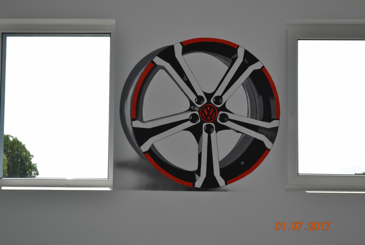 Iluzjonistyczne malowidło na ścianie przedstawiające felgę samochodową z logo Volkswagen, otoczone dwoma oknami, z datą 01.07.2017 w prawym dolnym rogu.