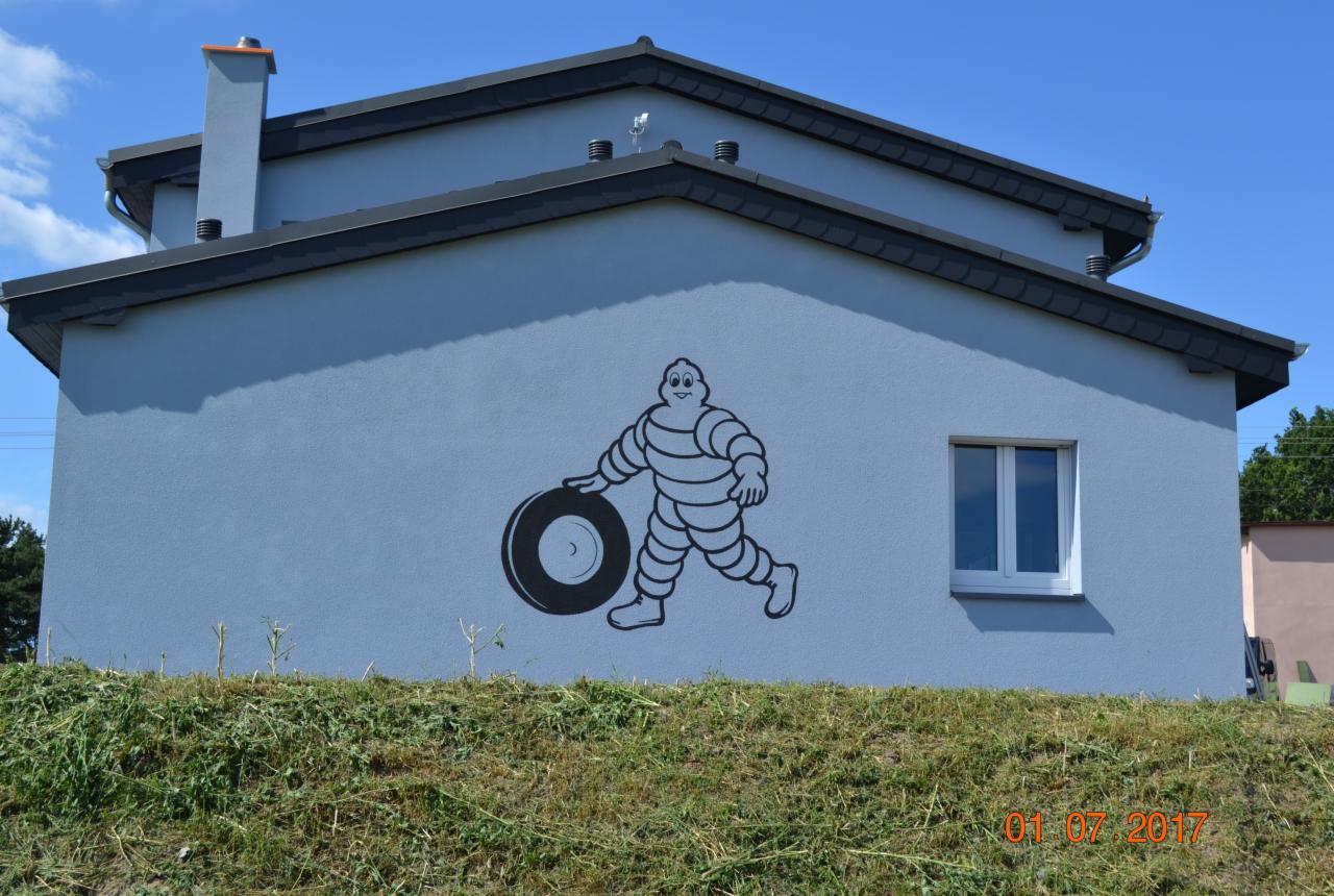 Jasnoniebieski dom z czarnym rysunkiem postaci z opony na elewacji, widok z poziomu trawy.